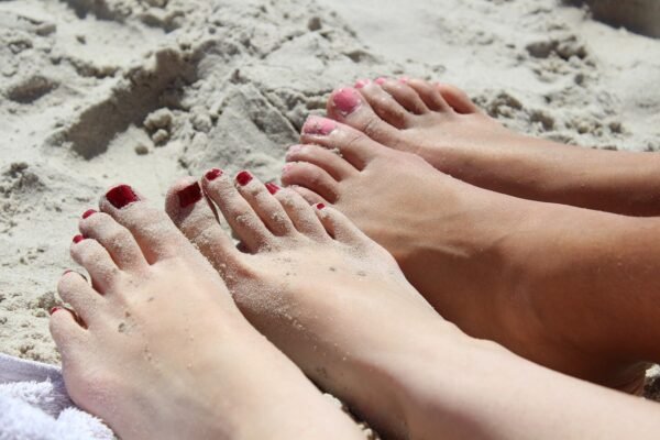 Two pairs of toes with painted nails in pink and red in the sand at the beach.